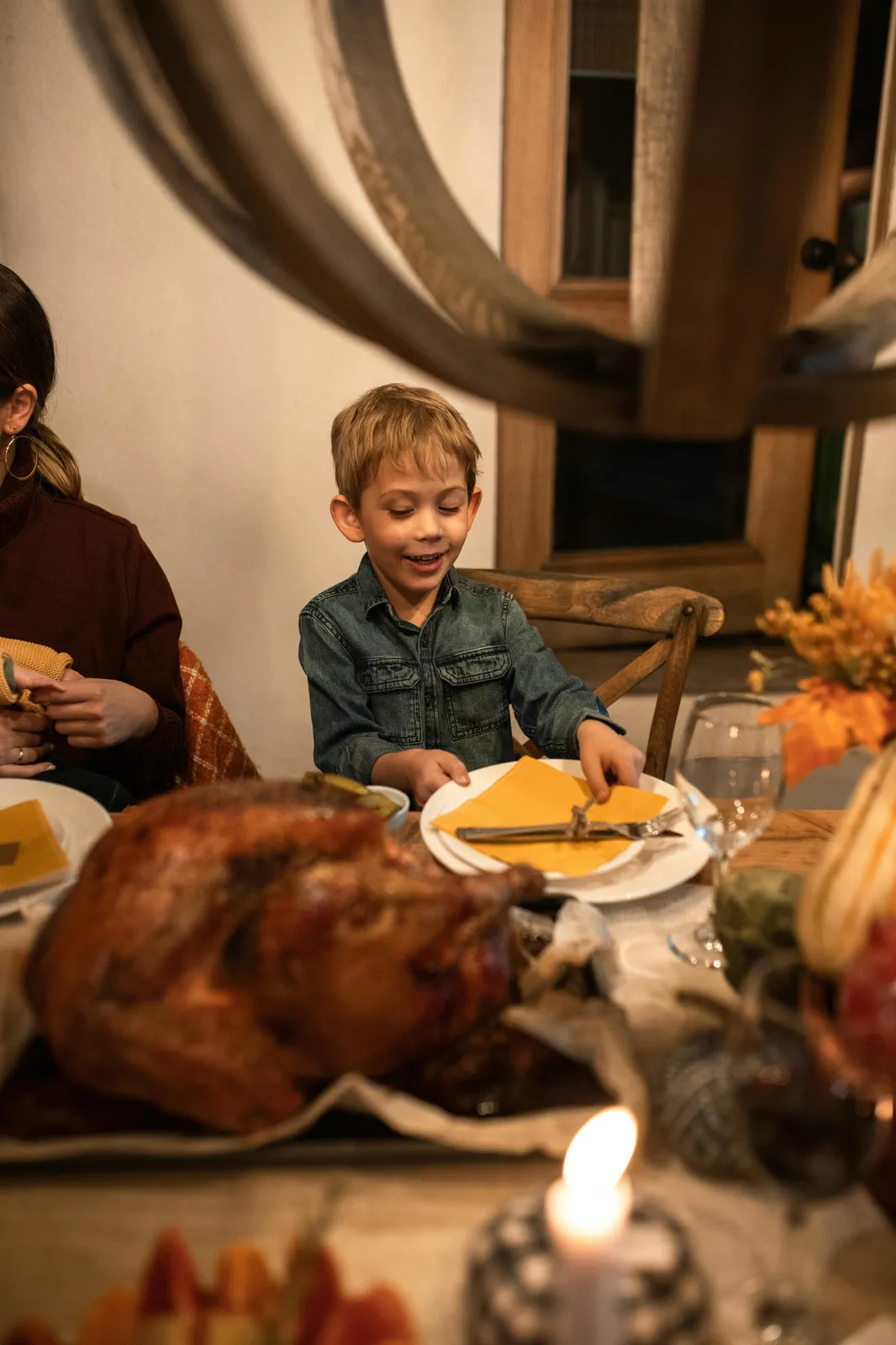 Niño sonriente sentado a la mesa de Acción de Gracias, con un pavo asado, velas y flores de otoño.