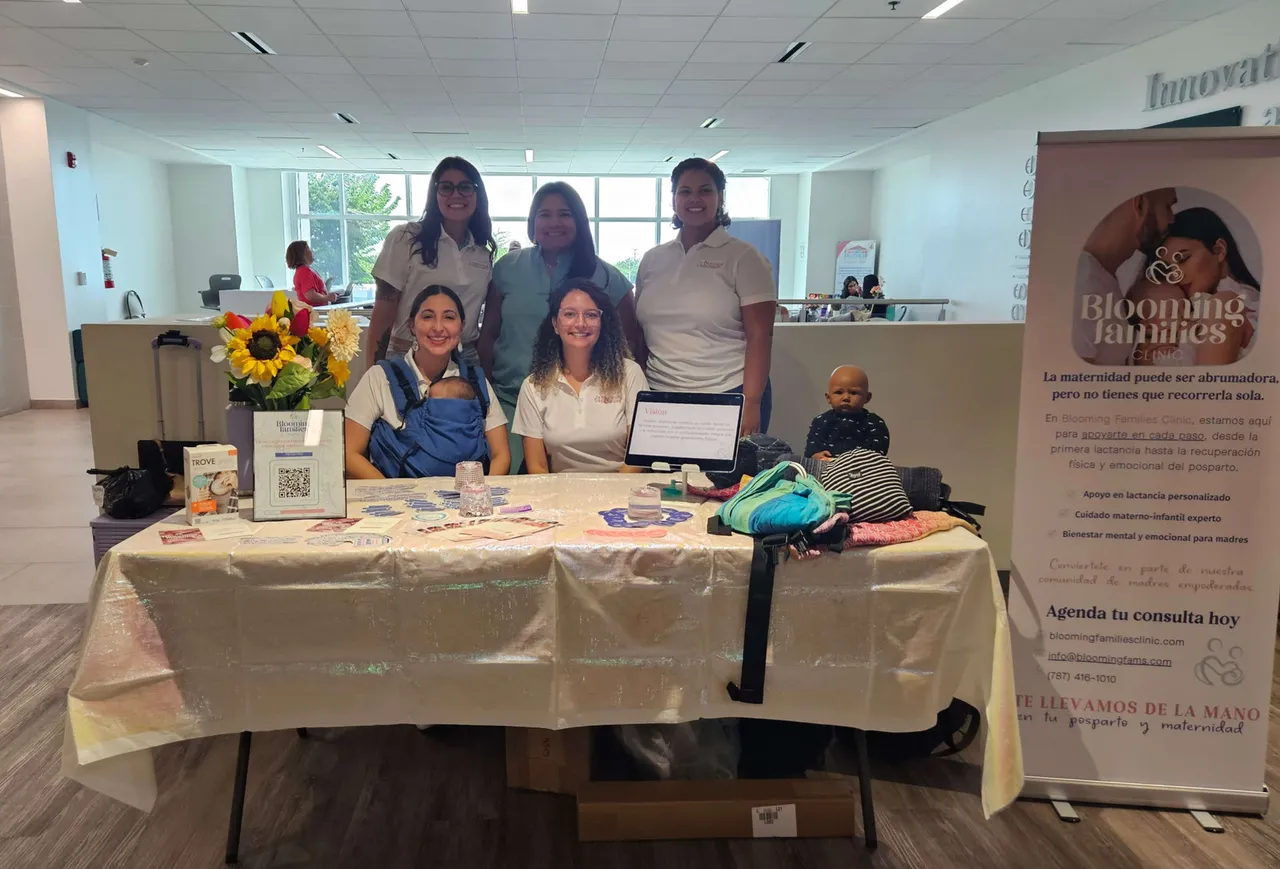 El equipo de Blooming Families Clinic posando junto a su mesa de exhibición en el MOMS Tour, mostrando recursos para el cuidado posparto y lactancia.